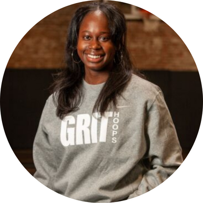 New York Grit Coach Cherkira Lashley, posing in their gray and white sweatshirt, one hand in pocket and smiling confidently against a basketball court backdrop for their official coach photo.