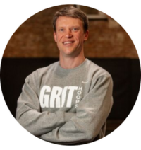 New York Grit Coach Brad Wetherell, posing in their gray and white sweatshirt, arms crossed and smiling confidently against a basketball court backdrop for their official coach photo.