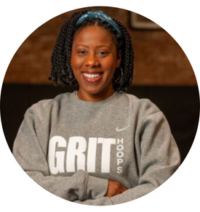 New York Grit Coach Camille Edwards, posing in their gray and white sweatshirt, arms crossed and smiling confidently against a basketball court backdrop for their official coach photo.